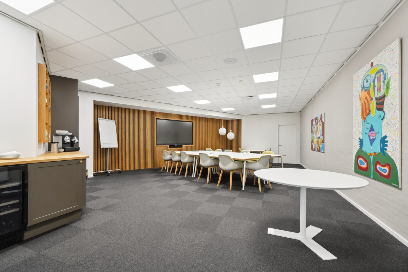 Moderne konferencelokale med langbord og stole, storskærm, flipover, skænk og farverige kunstværker på væggen. Lokalet har et neutralt farveskema og stærk belysning.