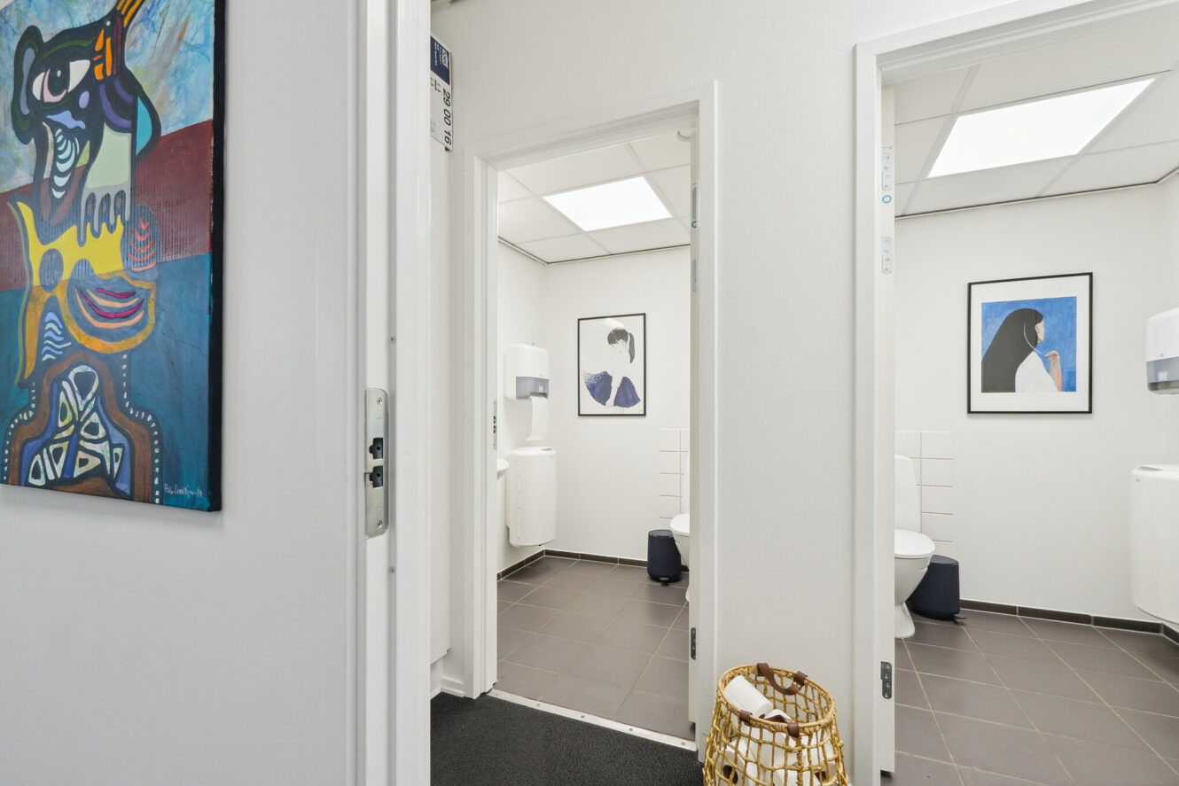 Hvid gang med to åbne badeværelsesdøre, moderne kunst på væggene, mørke klinkegulve og en flettet kurv i forgrunden.