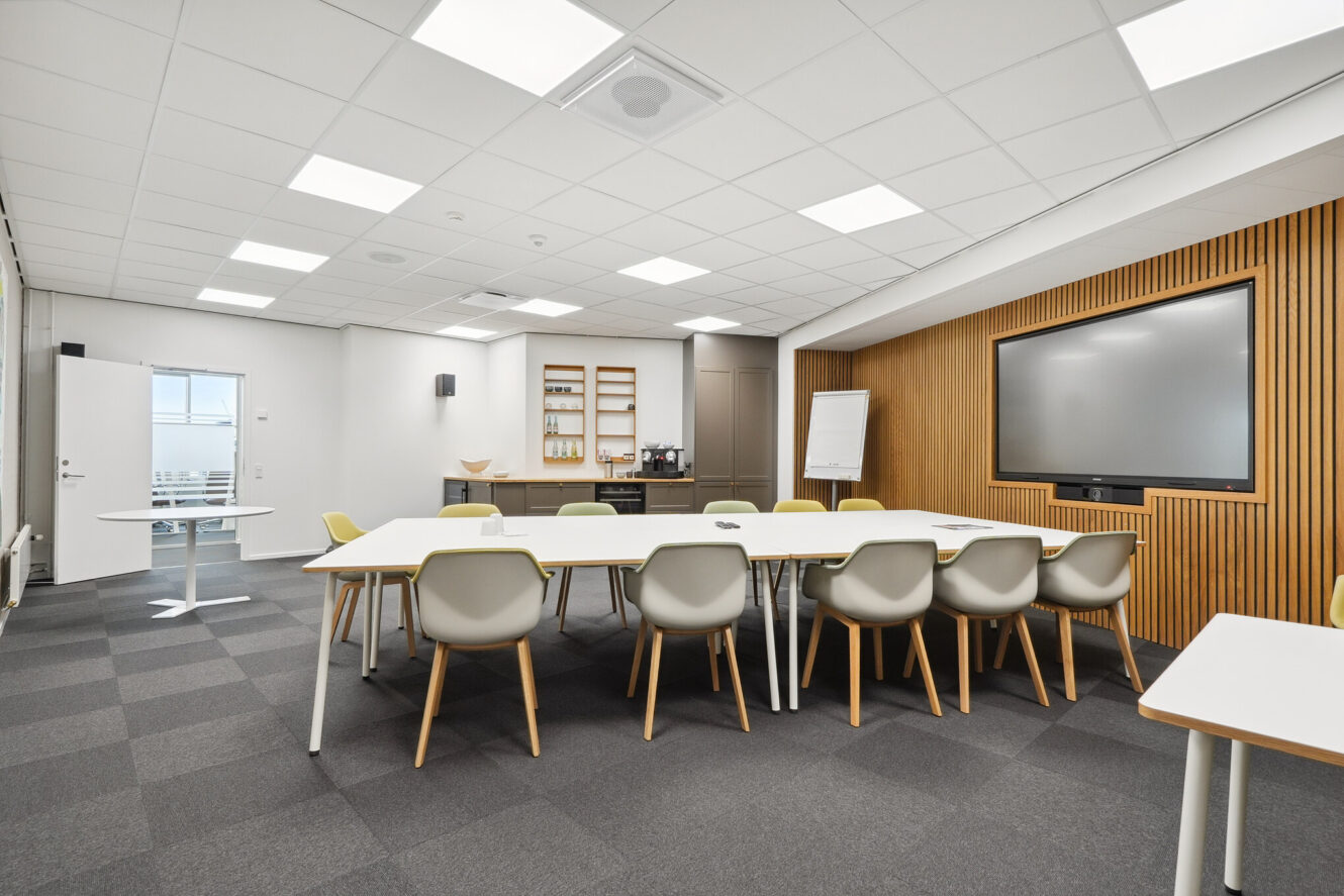 Moderne konferencelokale med hvide borde, beige stole, storskærm, flipover, vægreoler og loftslamper. Lokalet har gråt gulvtæppe og træpaneler.