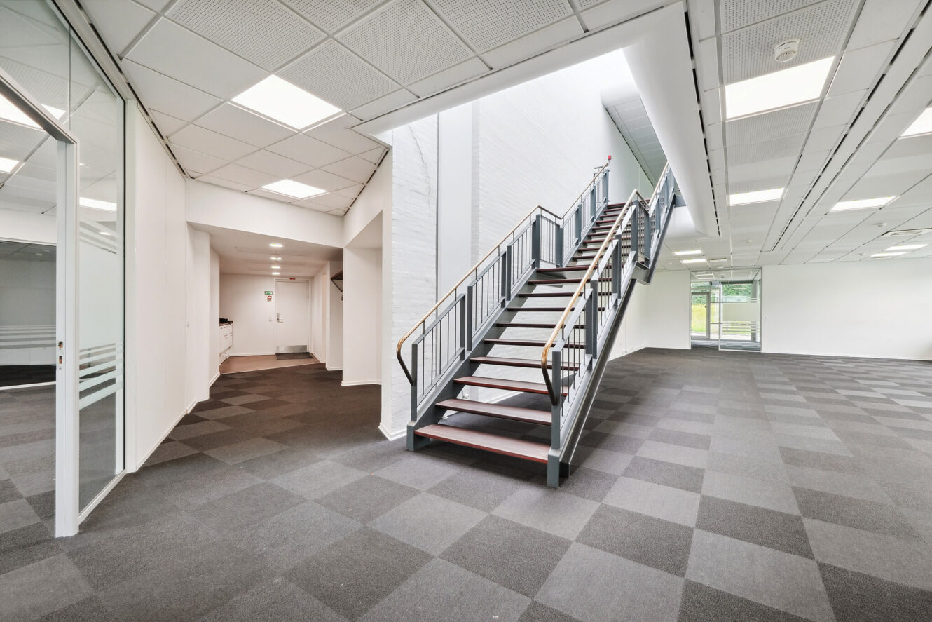 Moderne, tomt kontorinteriør med gråt gulvtæppe, hvide vægge, en trappe, der fører op til øverste etage, og store vinduer, der giver naturligt lys.
