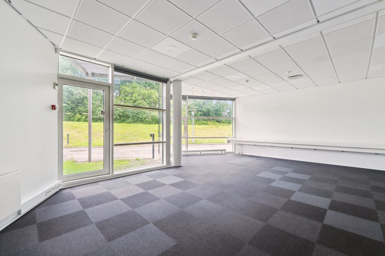 Tomt kontorlokale med ternet gulvtæppe, store vinduer, hvide vægge og udsigt til grønne områder udenfor.