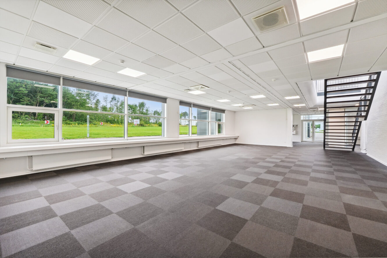 Rummeligt, tomt rum med store vinduer, ternet tæppe, hvide vægge og en sort trappe, der fører op til en overetage. Grøn græsplæne og træer er synlige udenfor.