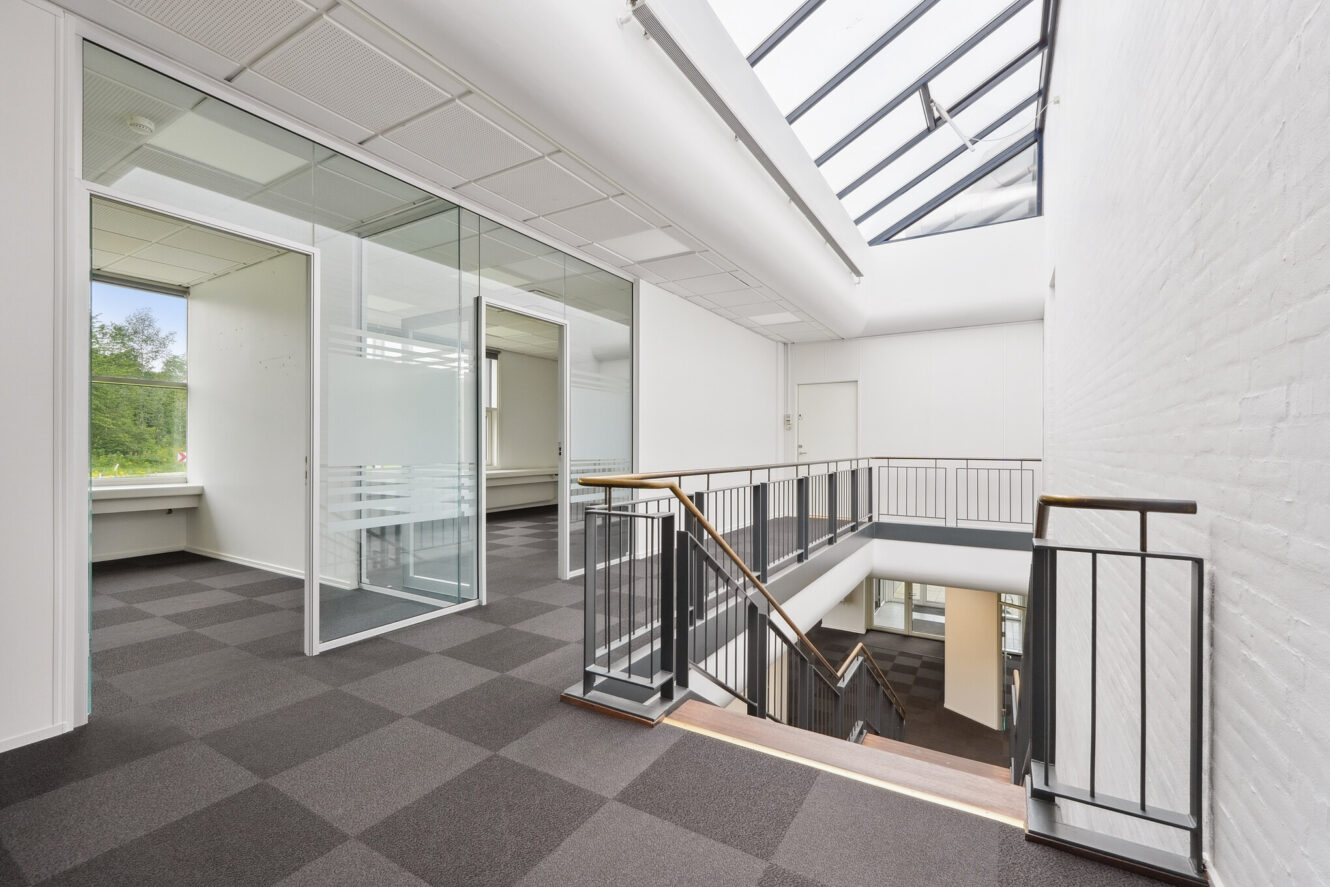 Moderne kontorgang med mødelokaler med glasvægge, brunternet gulvtæppe, trapperækværk i sort metal og et ovenlysvindue, der lukker naturligt lys ind.