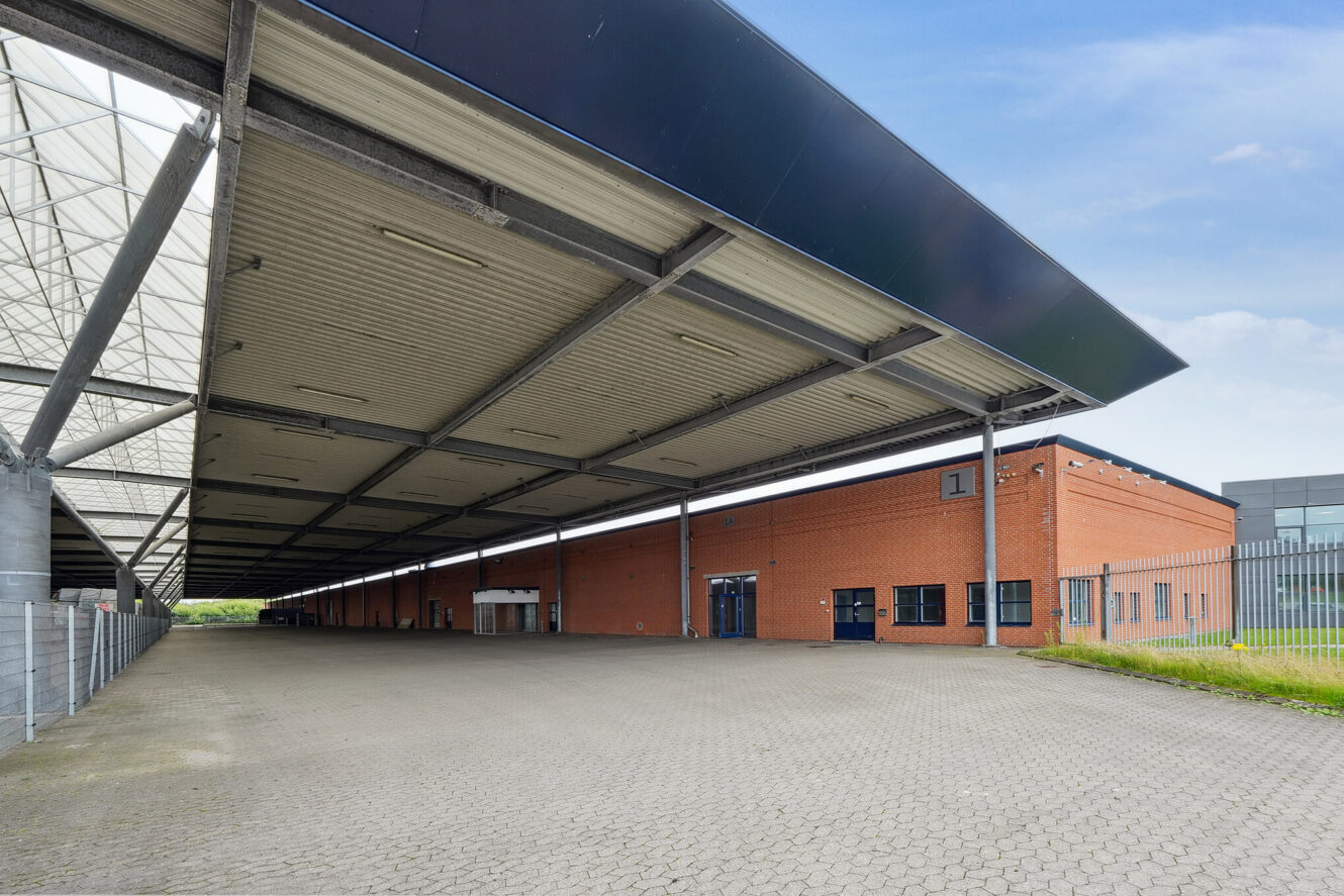 Stort overdækket læsseområde med en industribygning i mursten, metaltagkonstruktion og asfalteret jord under en blå himmel.
