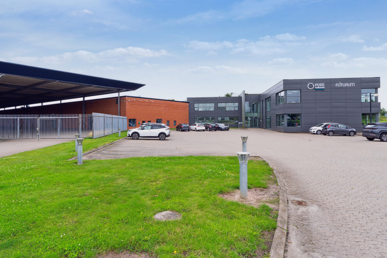 En moderne erhvervsbygning med en parkeringsplads, flere parkerede biler, grønt græs og en delvis overskyet himmel. Bygningen har skilte med PURE og ruhum.