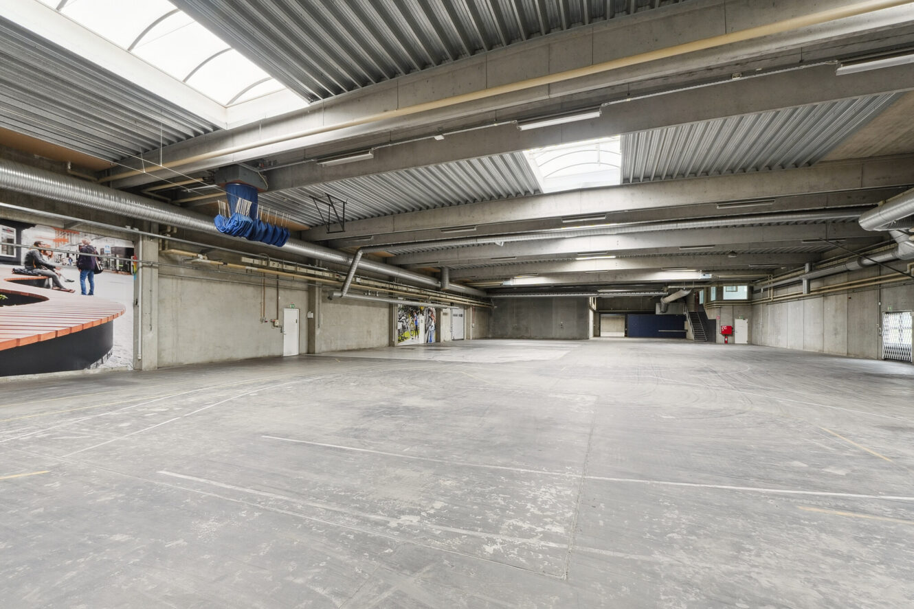 Stort tomt industrilager med betongulv, høje metallofter, ovenlys, ventilationskanaler og nogle vægmalerier eller plakater.