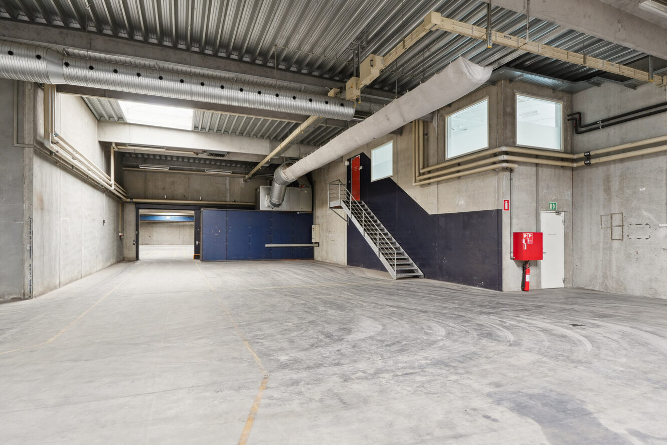 Et tomt industrilager med betonvægge, højt til loftet, luftkanaler, en metaltrappe, blå døre, store vinduer og en rød brandslukker monteret på væggen.