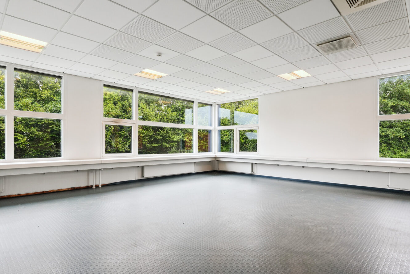 Tomt, umøbleret rum med store vinduer, hvide vægge og et gråt klinkegulv. Grønne træer er synlige udenfor gennem vinduerne. I loftet er der lysstofrør og ventilationsåbninger.
