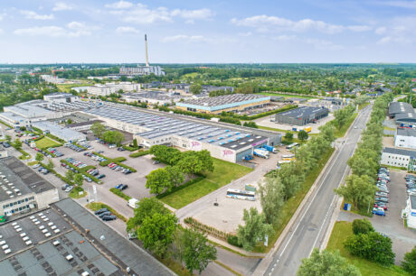 Luftfoto af et stort industrikompleks med flere lagerbygninger, parkerede køretøjer, grønne områder og omkringliggende veje under en delvis overskyet himmel.