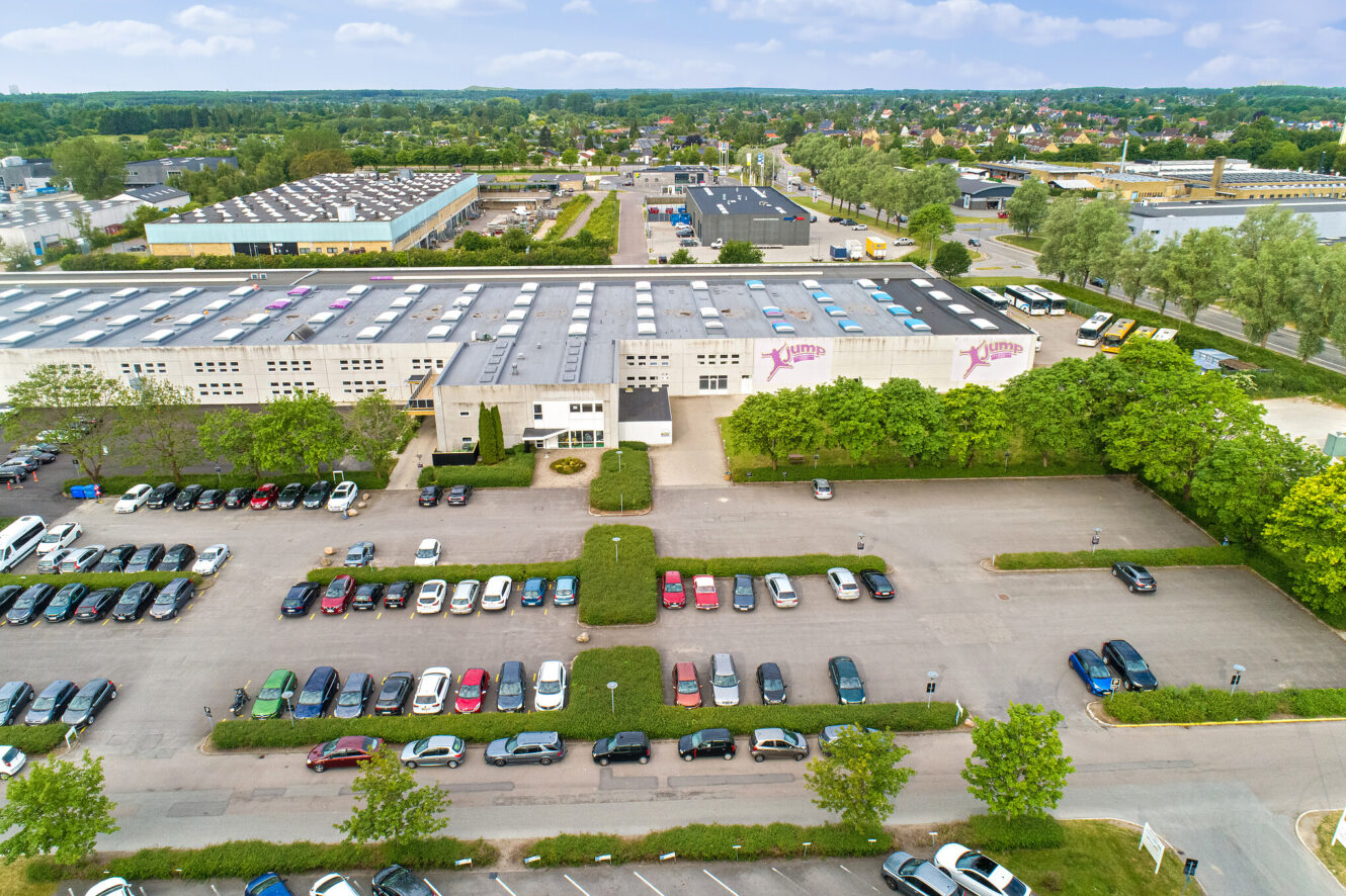 Luftfoto af en stor industribygning med tilstødende parkeringspladser og mange parkerede biler, omgivet af træer og andre bygninger i baggrunden.