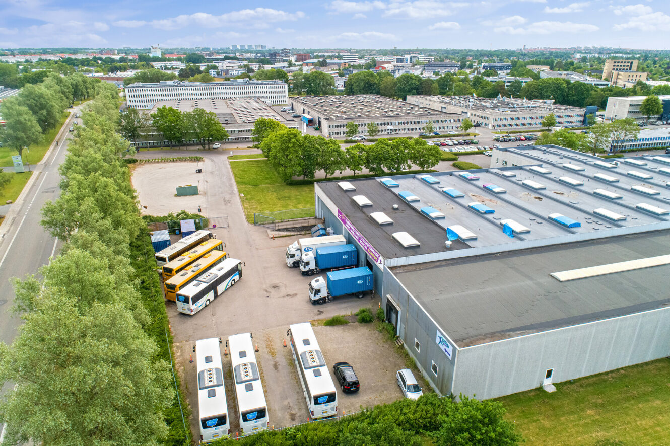 Luftfoto af et industriområde med lagerbygninger, parkerede busser, lastbiler og omgivende træer under en delvis overskyet himmel.