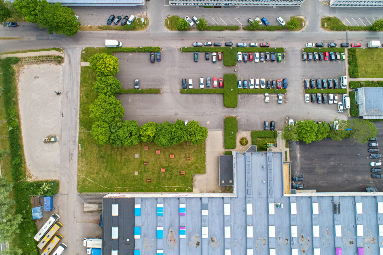 Luftfoto af en parkeringsplads med parkerede biler, tilstødende græsareal med træer og bænke og en del af en industribygning.