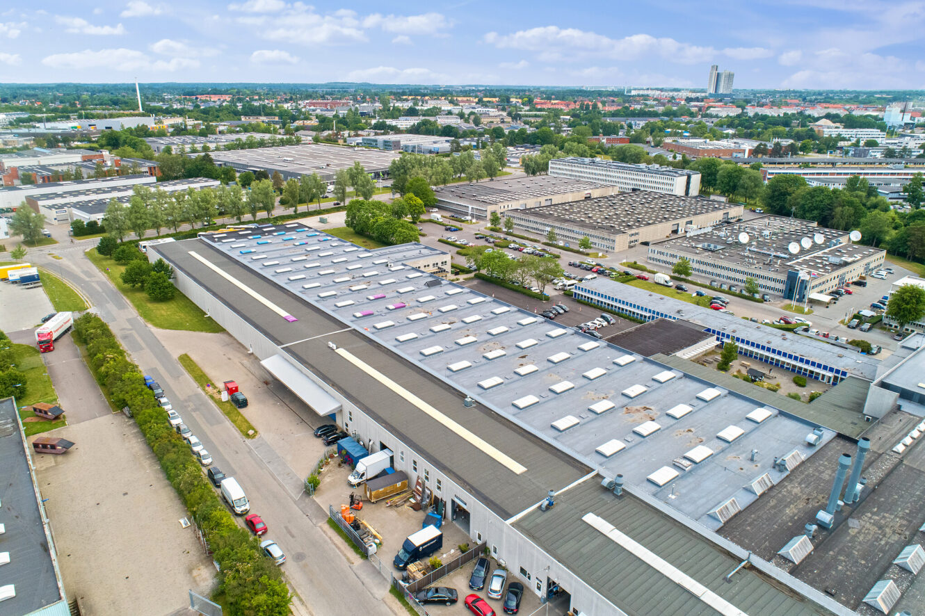 Luftfoto af et industrikompleks med store lagerbygninger, parkerede køretøjer, træer og et bybillede i baggrunden under en delvis overskyet himmel.