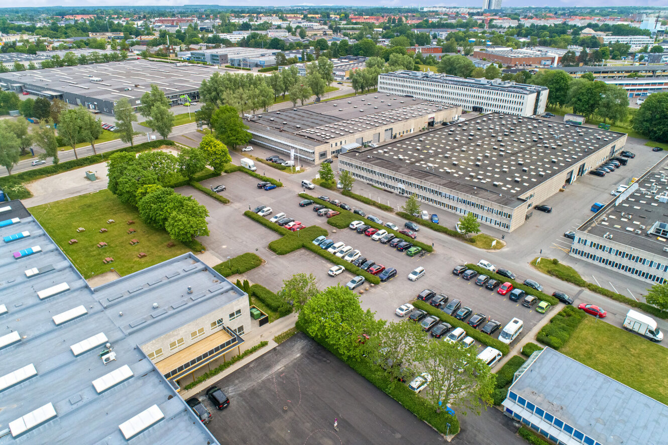 Luftfoto af en industripark med store lagerbygninger, parkeringspladser fyldt med biler og grønne områder med træer og græs.