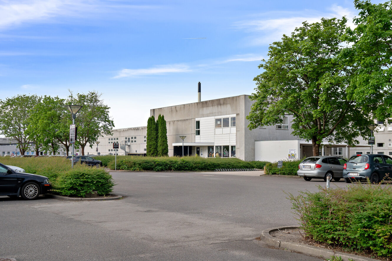 En parkeringsplads med flere parkerede biler foran en moderne, grå betonbygning omgivet af træer under en delvis overskyet himmel.