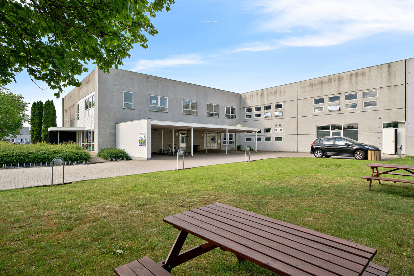 En moderne betonbygning i to etager med flere vinduer, et lille indgangsparti, en parkeret sort bil og picnicborde af træ på en græsplæne.