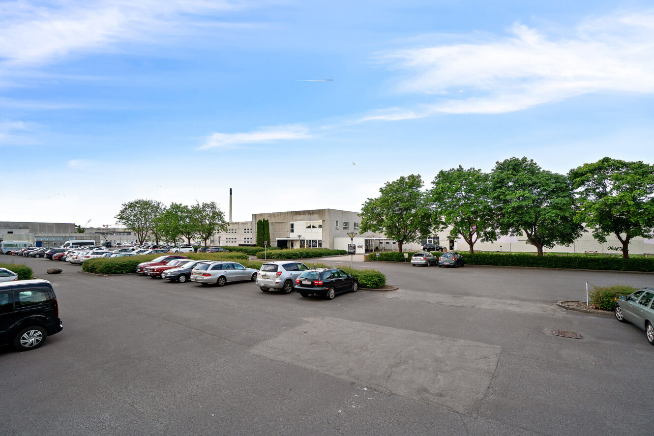En parkeringsplads med flere parkerede biler foran en stor, hvid industri- eller kontorbygning, omgivet af grønne træer under en delvis overskyet himmel.