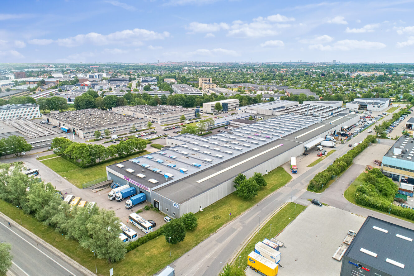 Luftfoto af et stort industrielt lagerkompleks med læsseramper, parkerede lastbiler og omkringliggende grønne områder i et bymiljø.