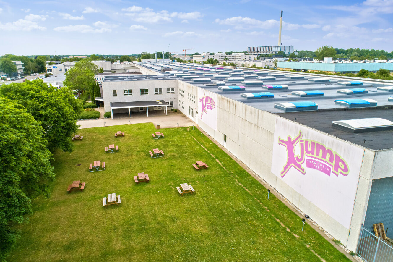 En stor hvid bygning med Jump-skiltning, omgivet af græs, træer og picnicborde, med et industriområde og en skorsten i baggrunden under en blå himmel.