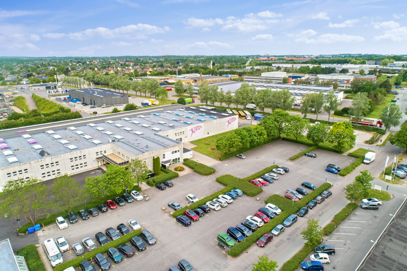 Luftfoto af en stor erhvervs- eller industribygning med en parkeringsplads fyldt med biler, omgivet af træer og andre bygninger under en delvis overskyet himmel.