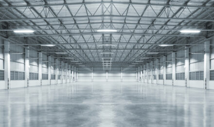 Interiør i en stor, tom lagerbygning med polerede betongulve, højt til loftet og synlige metalbjælker, stærkt oplyst af loftslamper.