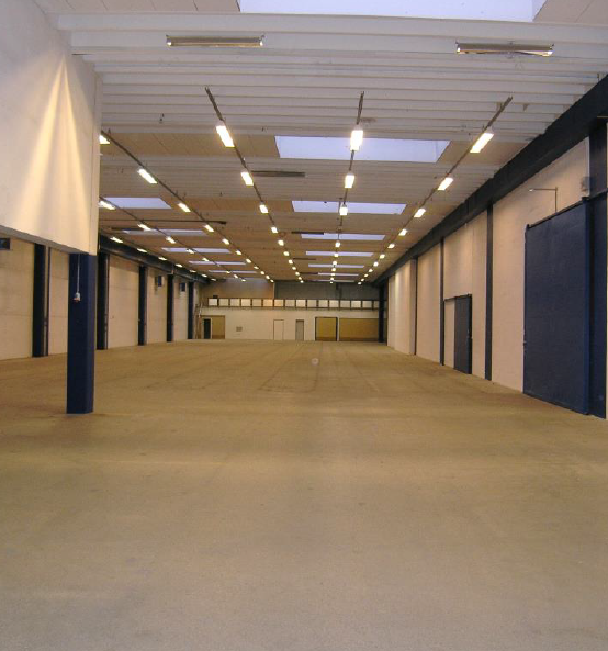 Stor tom lagerhal med højt til loftet, ovenlys, lysstofrør og glatte vægge. Betongulvet strækker sig til bagsiden, hvor flere døre og vinduer er synlige.