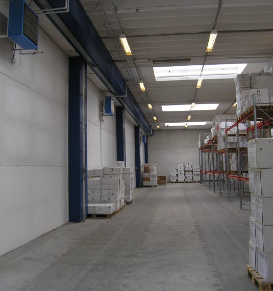 Et lagerinteriør med stablede kasser på metalreoler og paller langs betonvægge, oplyst af lysstofrør og ovenlysarmaturer.