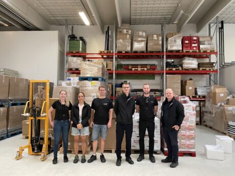 Syv personer står i en lagerhal foran hylder fyldt med kasser og forsyninger, og til venstre ses en gul palleløfter.