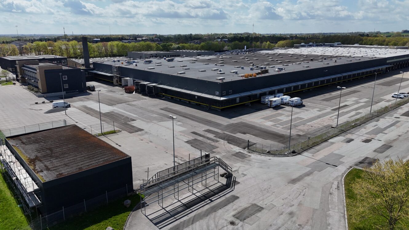 En stor, sort industriel lagerbygning med flere lastbiler parkeret udenfor, omgivet af en stort set tom parkeringsplads og indhegnet område på en solrig dag.