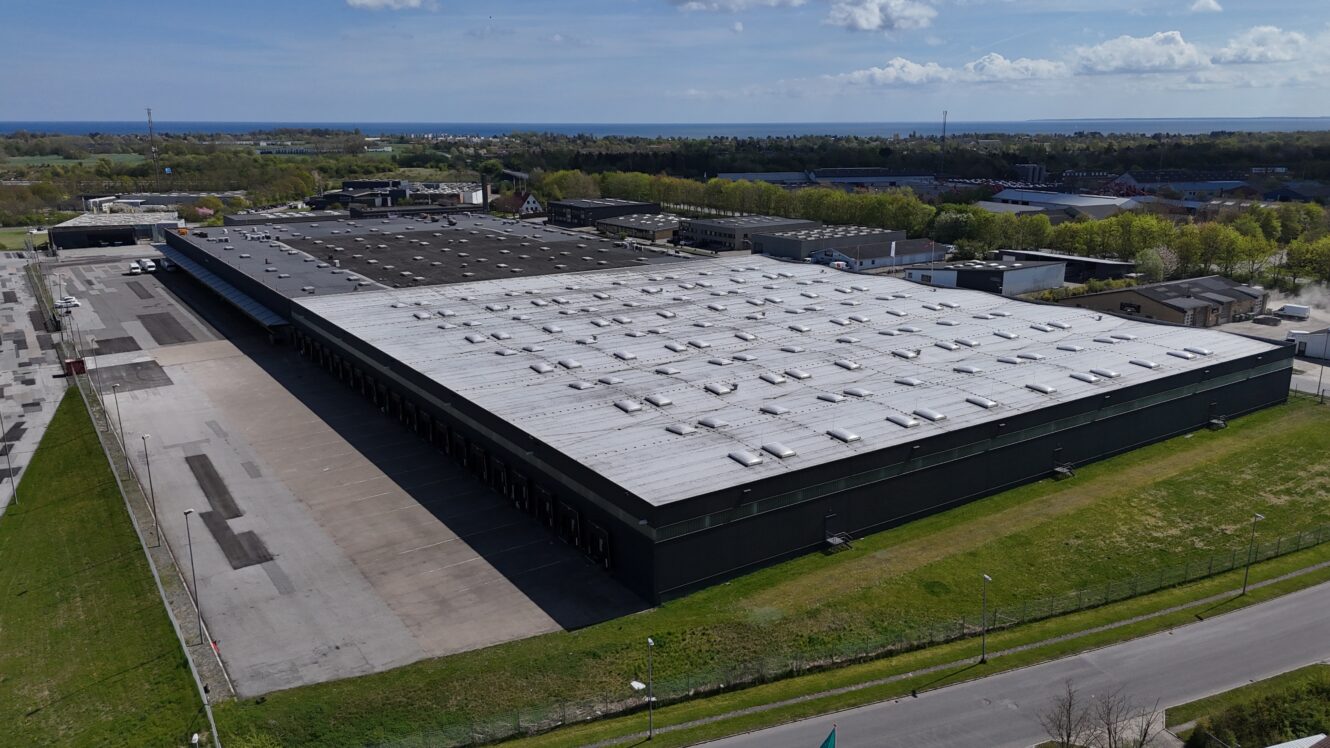 Et stort, rektangulært industrilager med hvidt tag er omgivet af asfalterede områder og grønne områder, og i baggrunden kan man se flere bygninger og en kystlinje.