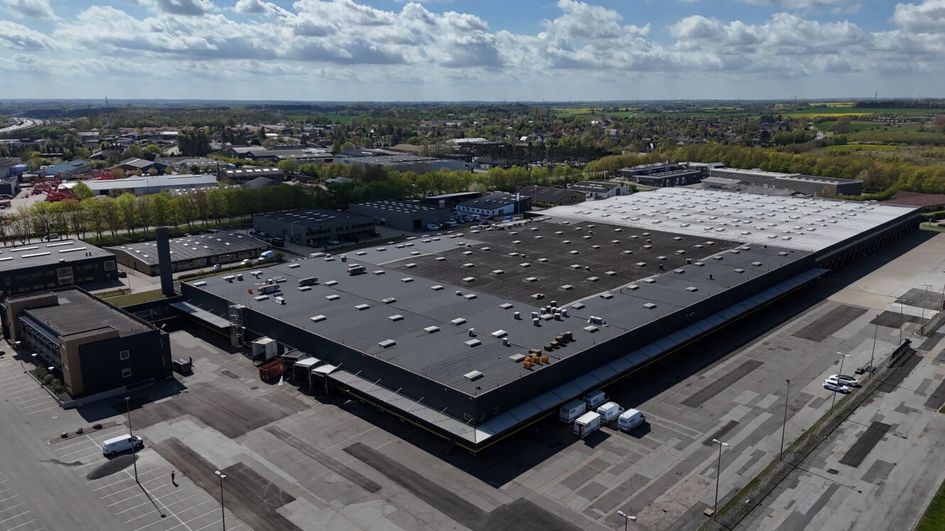 En stor lagerbygning med fladt tag og flere læsseramper og lastbiler ligger i et industriområde, omgivet af parkeringspladser og grønne områder under en delvis overskyet himmel.