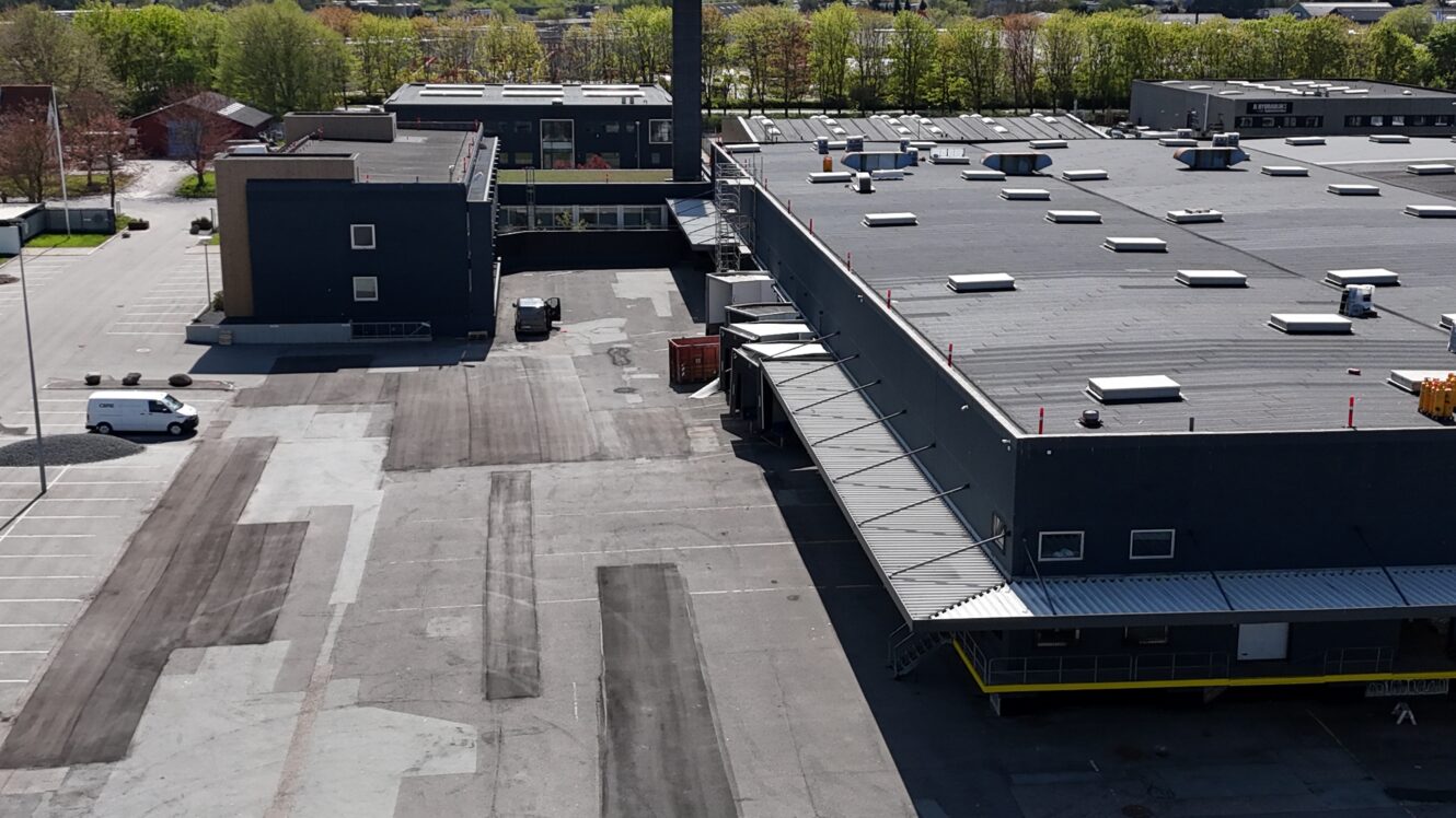 Luftfoto af en industribygning med et stort fladt tag, læsseramper, tilstødende parkeringsområder og omkringliggende grønne områder i baggrunden.