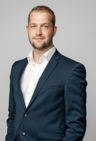 En mand med kort hår og skæg iført hvid skjorte og mørkeblå habitjakke står på en almindelig lysegrå baggrund med ansigtet mod kameraet og smiler let.