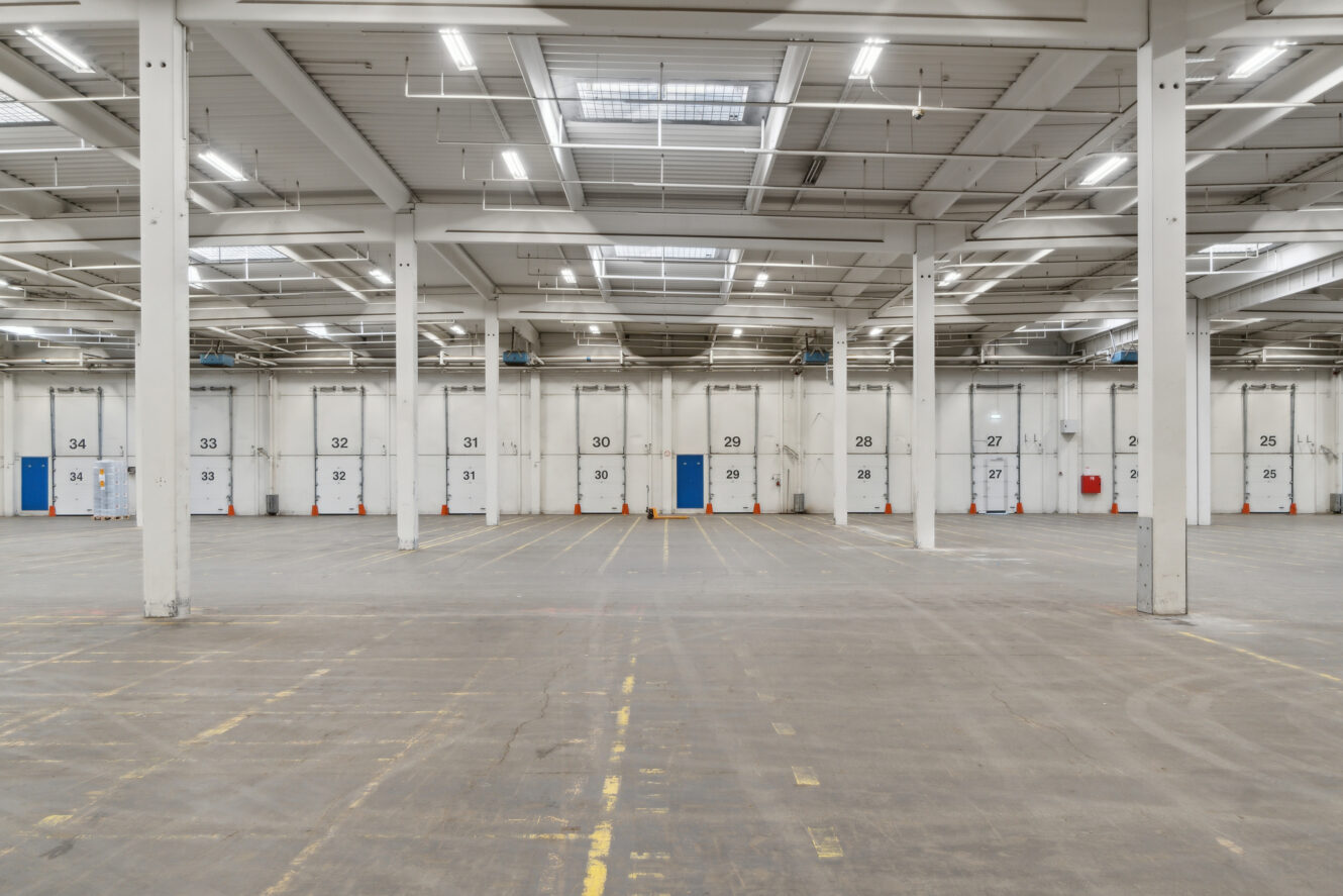 Stor, tom lagerhal med nummererede, hvide porte til læsseramper og betongulv, oplyst af lysstofrør og ovenlyspaneler i loftet.