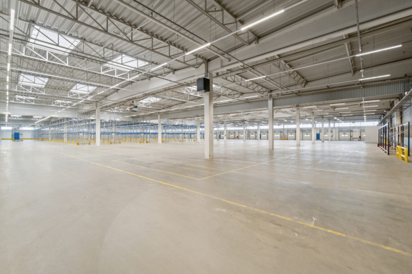 Rummeligt, tomt industrielt lagerinteriør med højt til loftet, synlige bjælker, ovenlys og betongulv. Rækker af metalreoler langs den ene side.