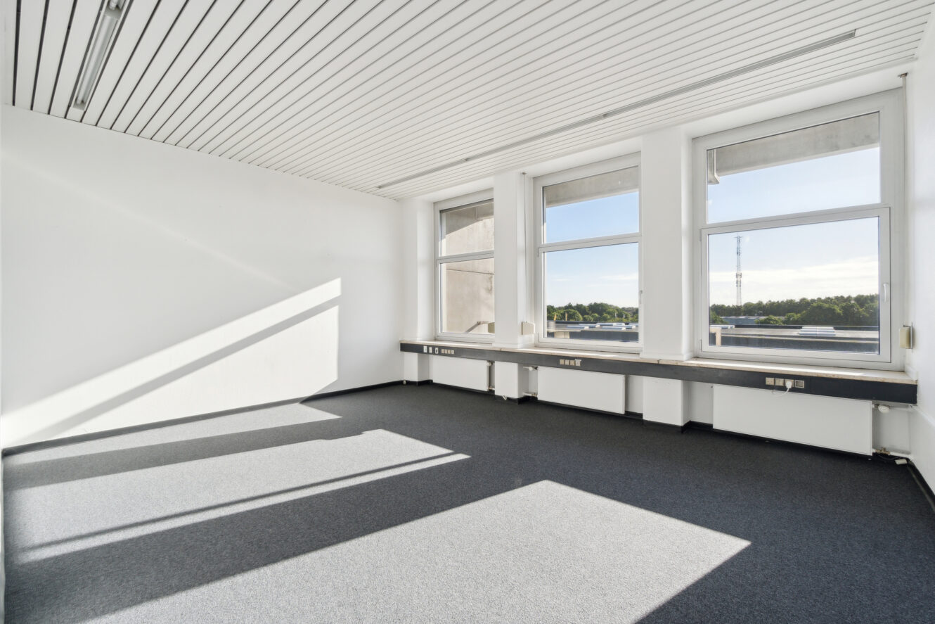 Tomt kontorlokale med store vinduer, hvide vægge, gulvtæppe og indbyggede skriveborde langs vinduerne. Sollyset strømmer ind og kaster skygger på gulvet.