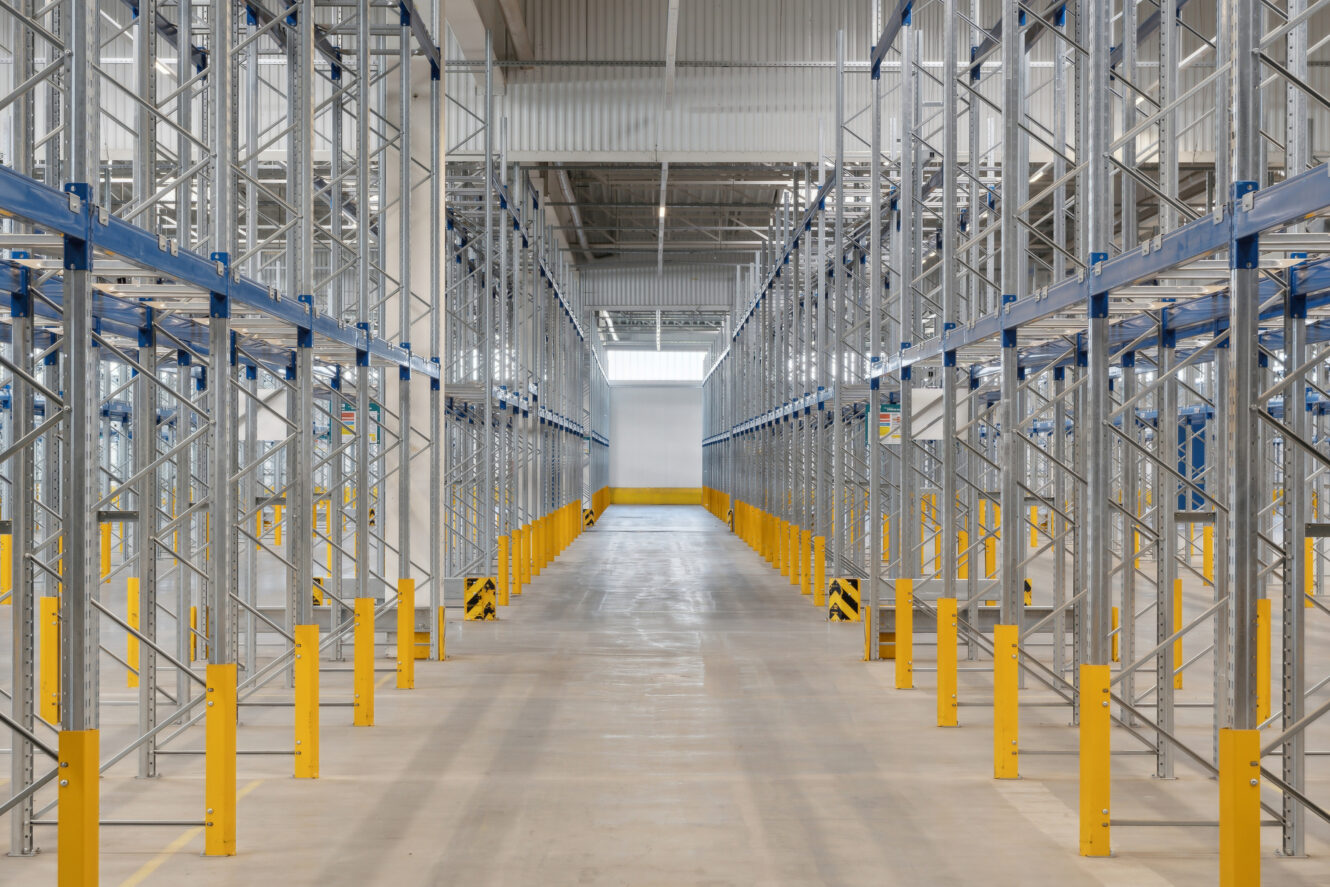 Tomt lagerinteriør med høje metalreoler, gule sikkerhedsbarrierer og en bred midtergang under et højt loft.