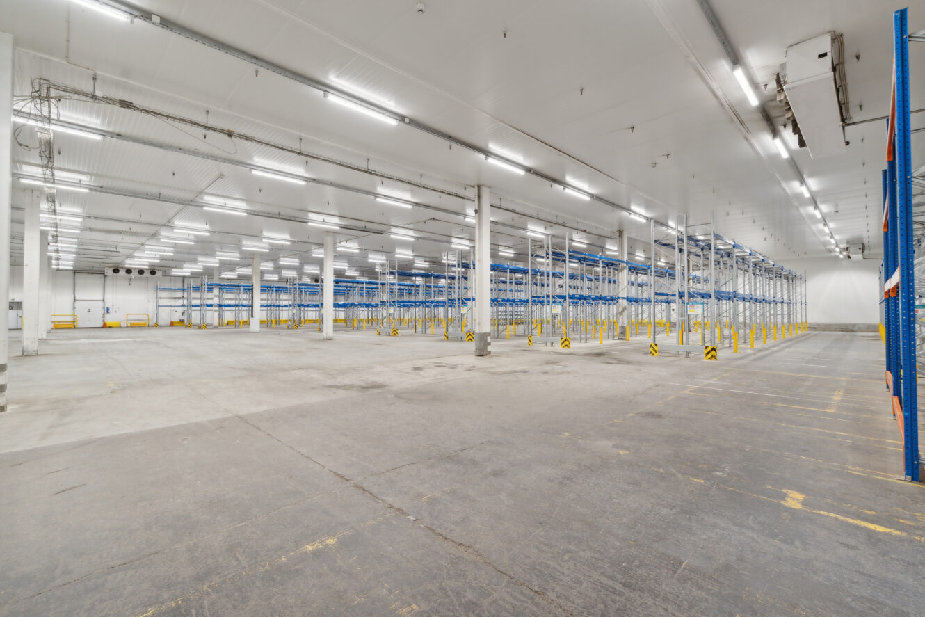 En stor, tom lagerhal med hvide vægge, højt til loftet og blå metalreoler i rækker.
