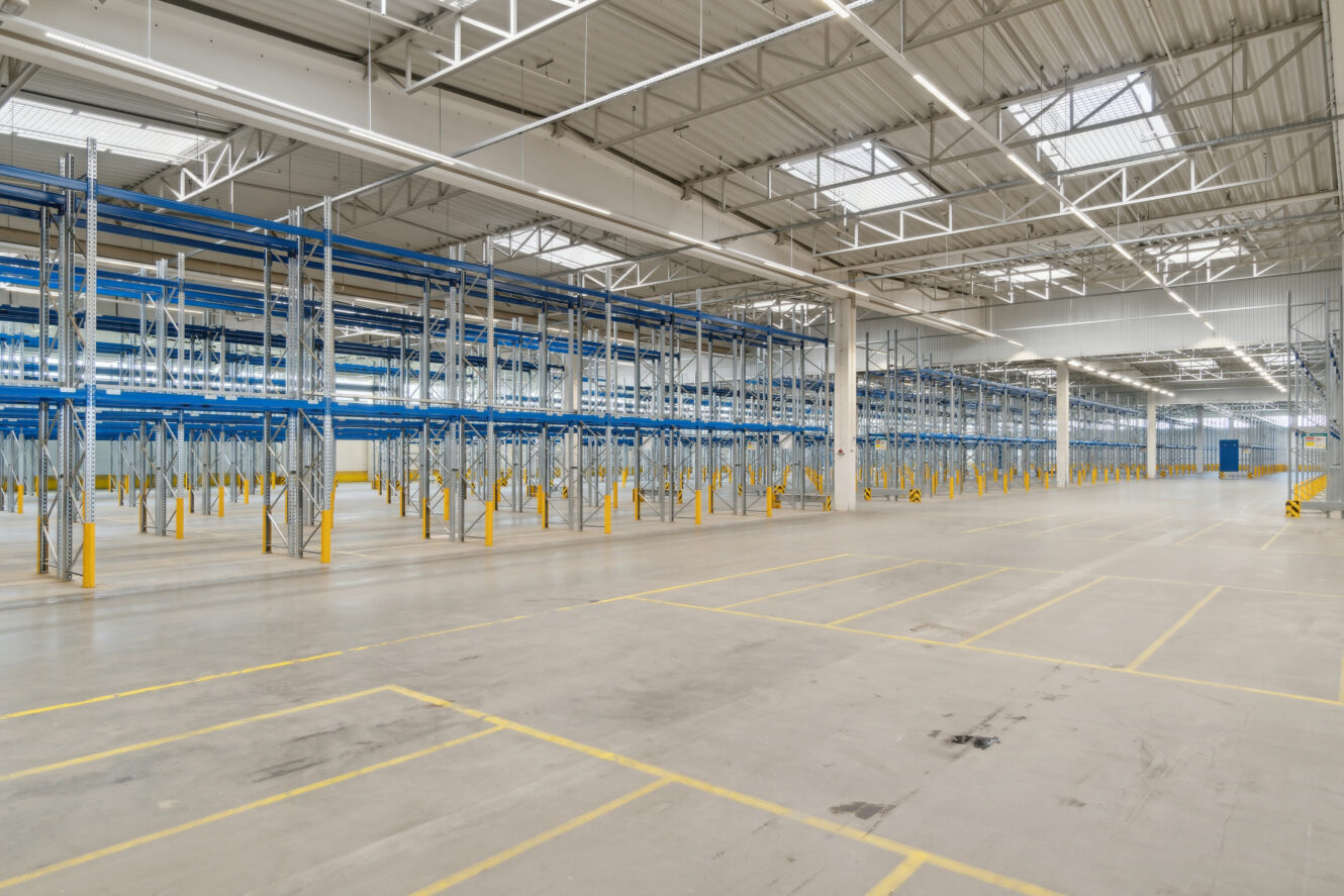 Tomt lagerinteriør med højt til loftet, lagerreoler i metal og gule gulvmarkeringer under kraftig overbelysning.