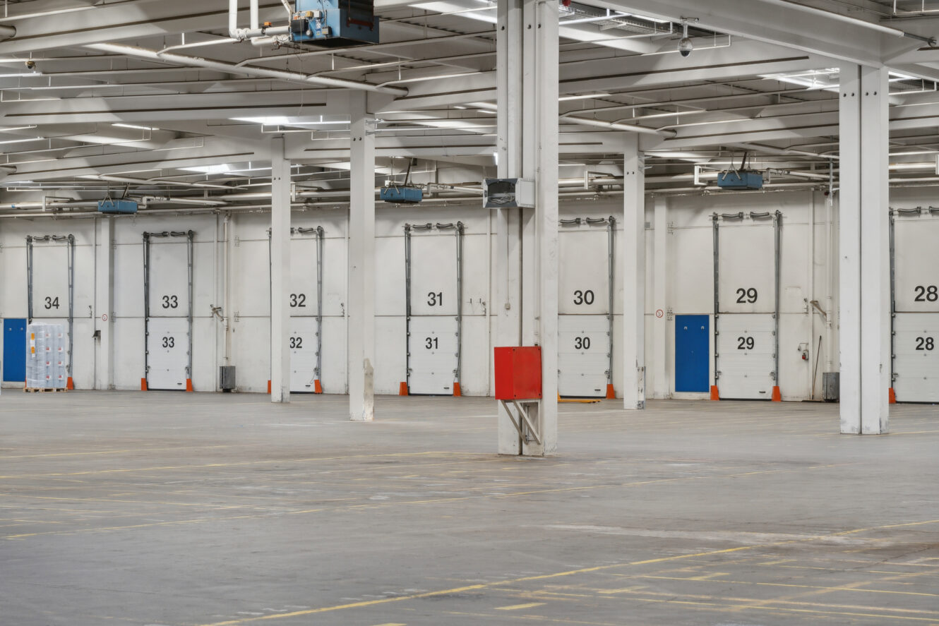 Interiør af en tom lagerbygning med nummererede læsseramper, hvide vægge, synligt loft og en rød brandsikringskasse monteret på en søjle.