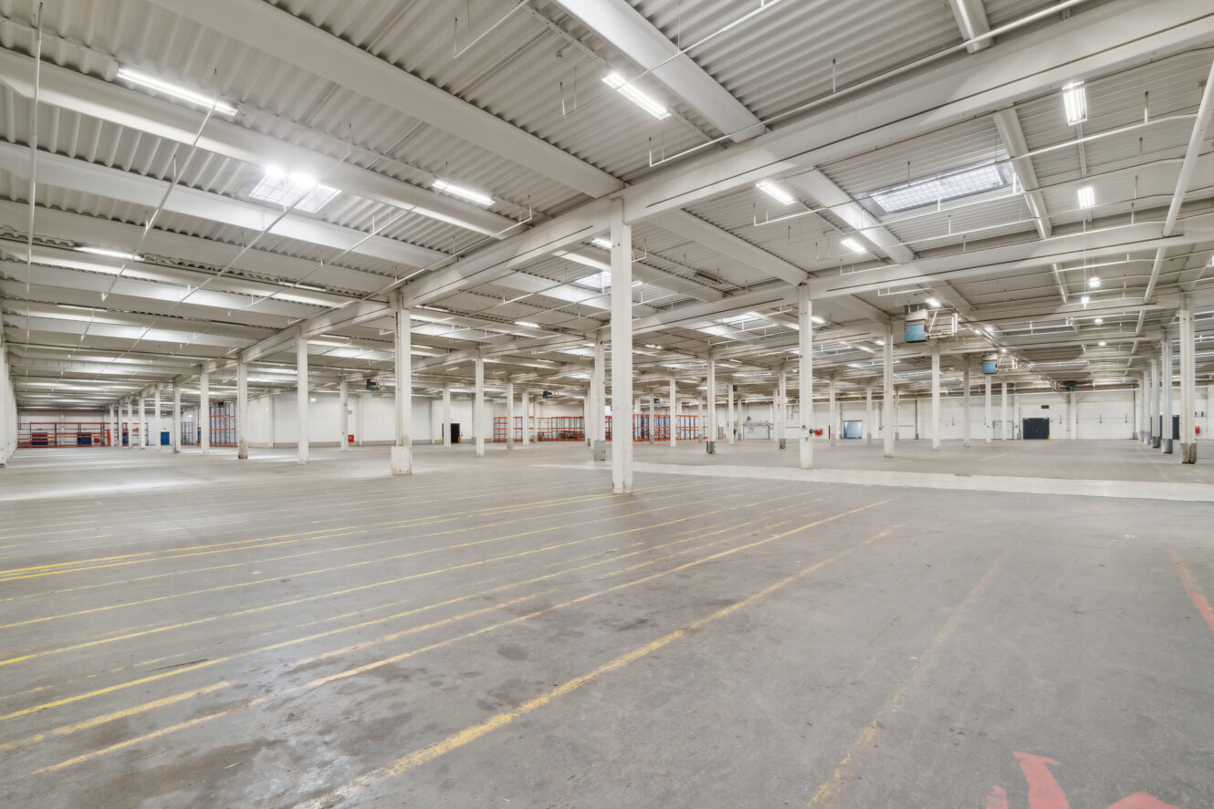 Stort, tomt lagerinteriør med højt til loftet, ovenlys, hvide vægge og markerede linjer på betongulvet.