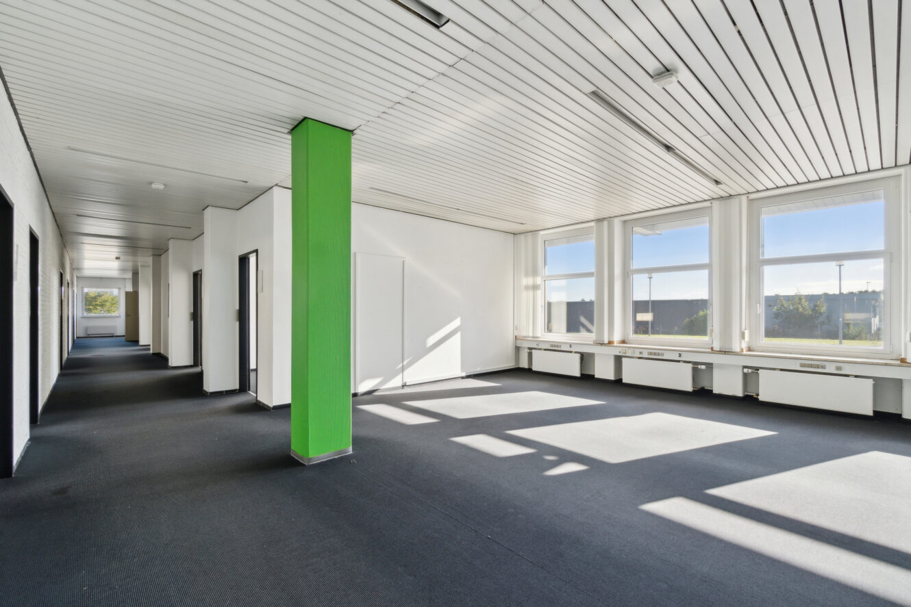 Tomt kontorlokale med hvide vægge, en grøn søjle, store vinduer, der lukker naturligt lys ind, og et mørkt gulvtæppe.