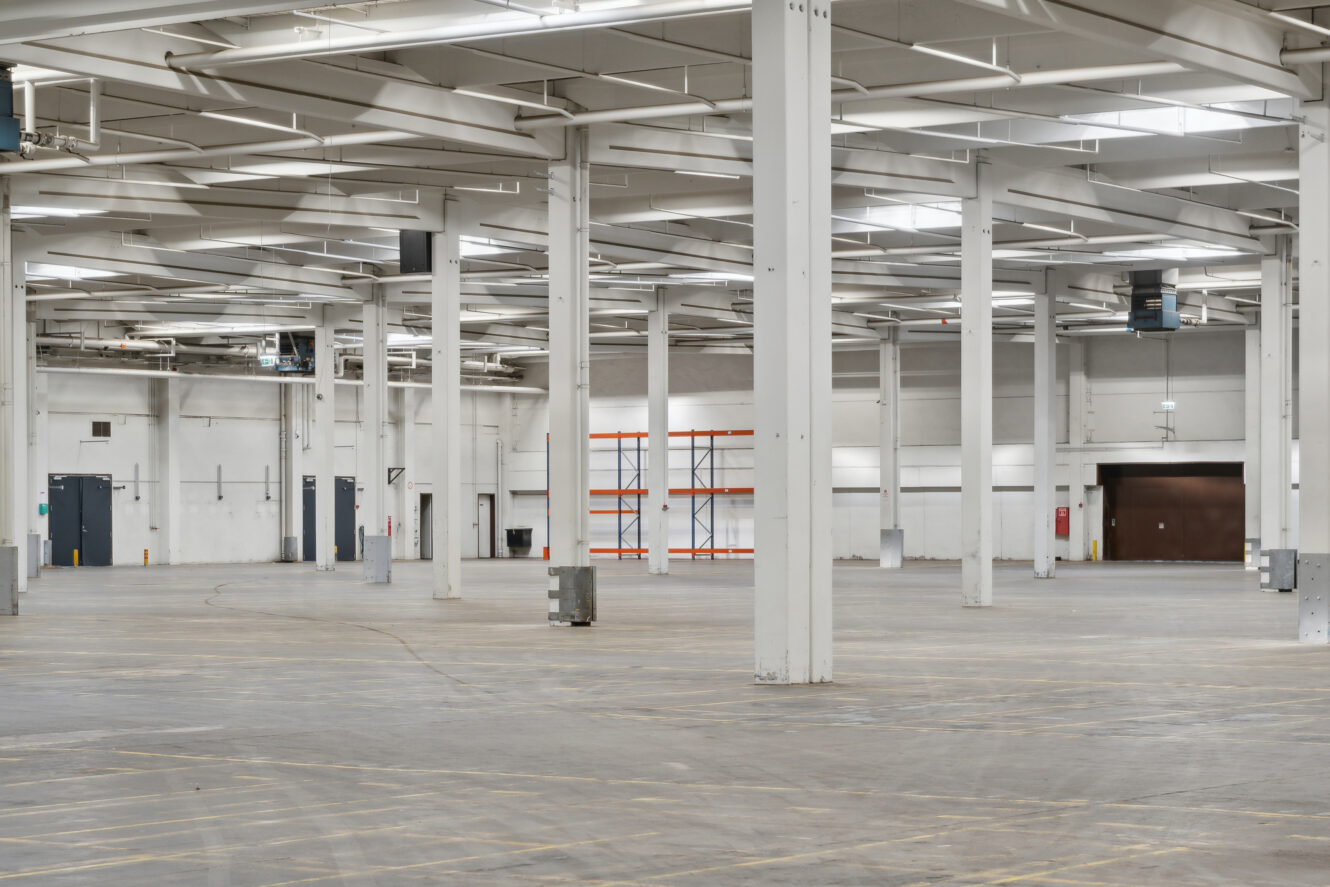 Stort, tomt industrilager indvendigt med betongulv, hvide støttesøjler og ovenlys. Reoler og porte til læsseramper er synlige i baggrunden.