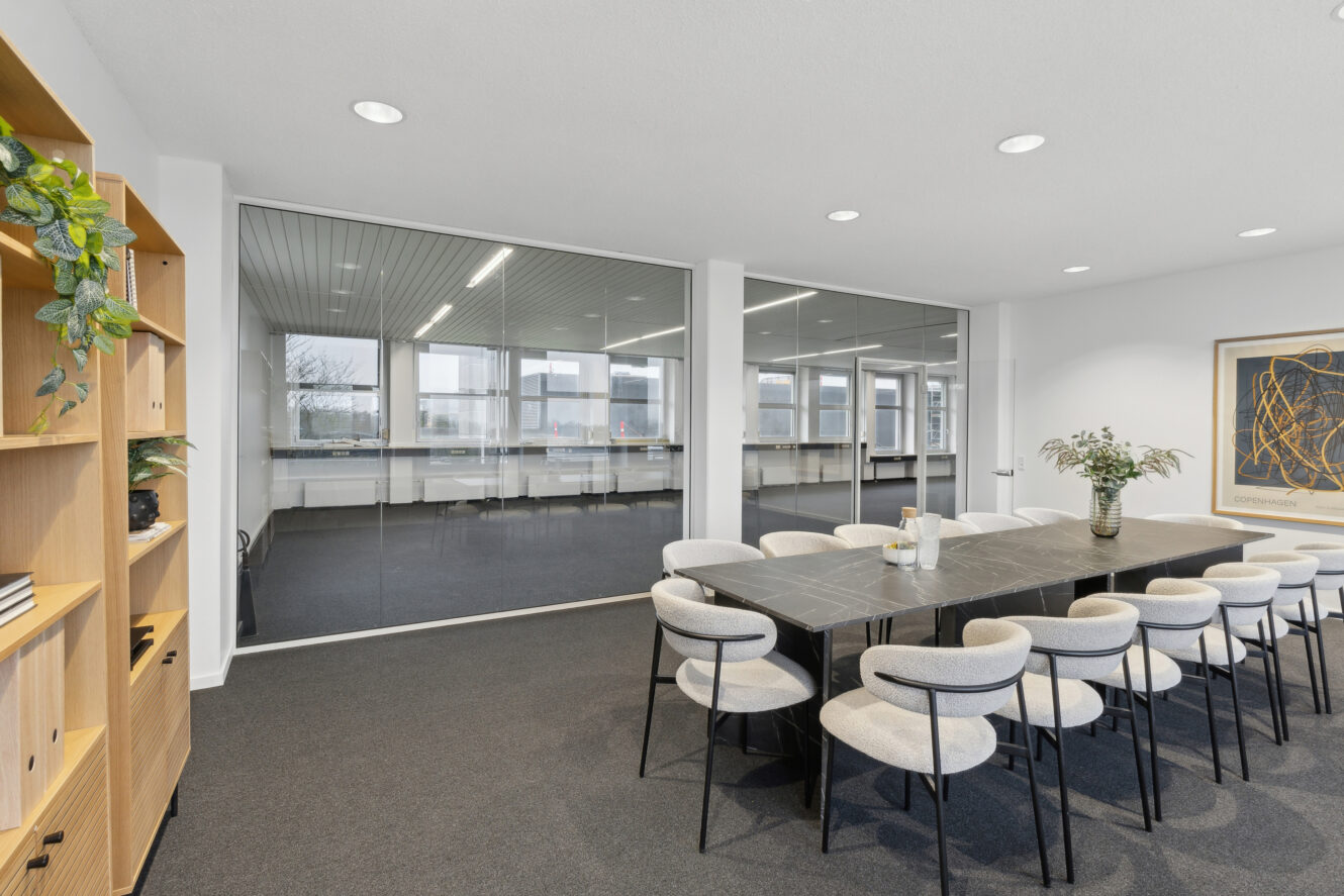 Moderne konferencerum med et langt sort bord, tolv hvide stole, en potteplante, hylder med bøger og glasvægge, der afslører et andet kontor.