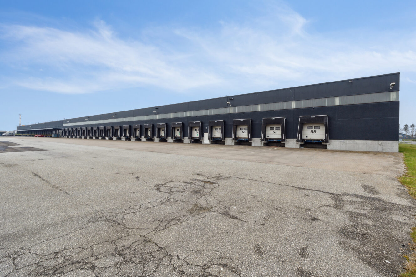Et bredt udsyn over et industrilager med flere læsseramper, hver markeret med et nummer, under en delvis overskyet himmel.