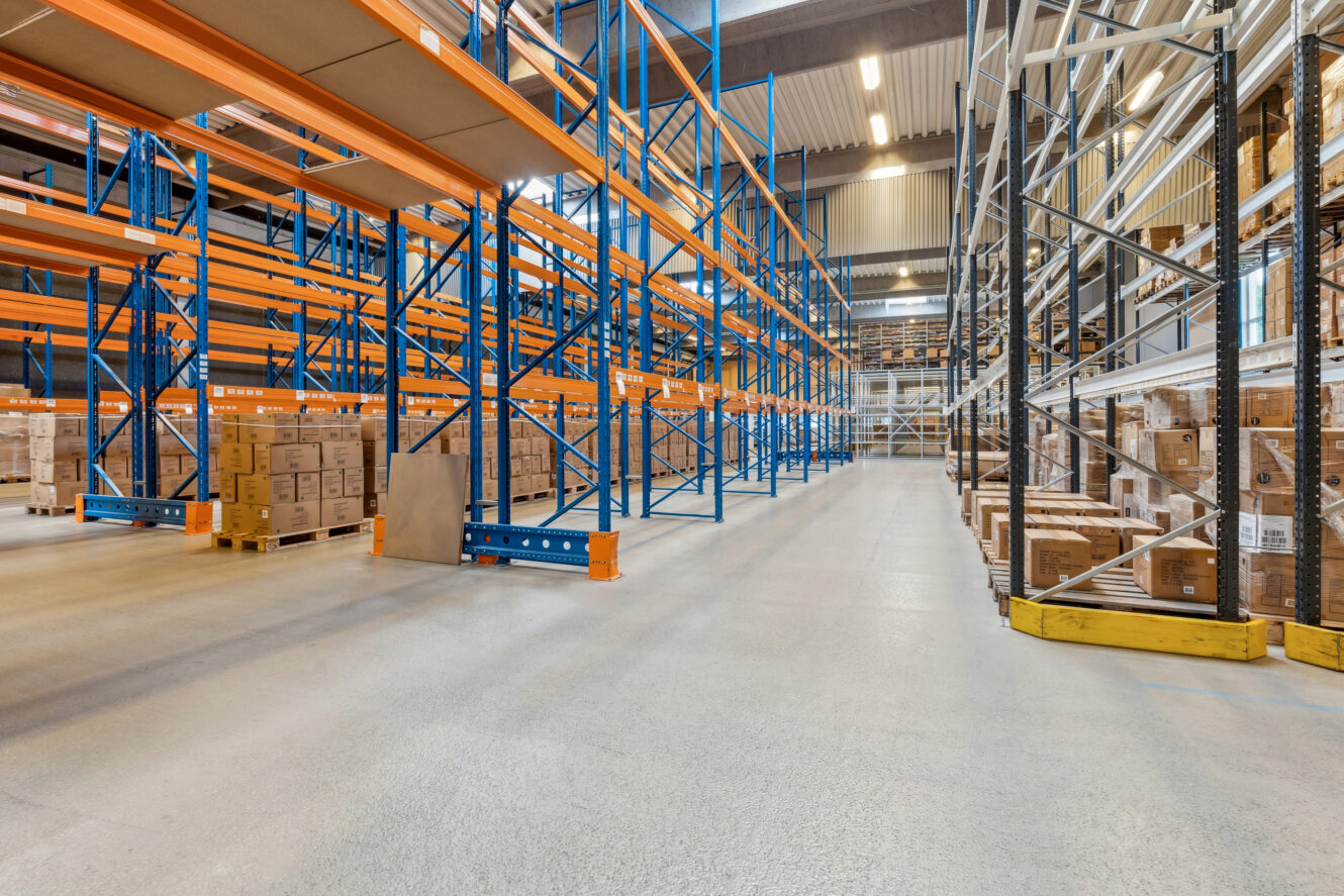 Stort lagerinteriør med høje blå og orange reoler, hvoraf nogle er fyldt med papkasser, og brede, rene gange.