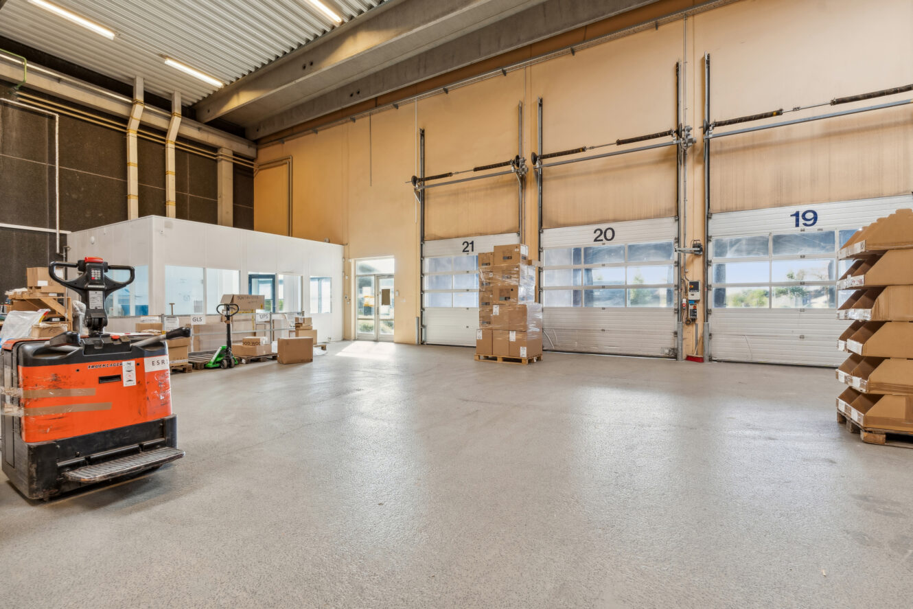 Rummeligt lagerinteriør med stablede papkasser, palleløfter og store læsseramper med numrene 19 til 21.