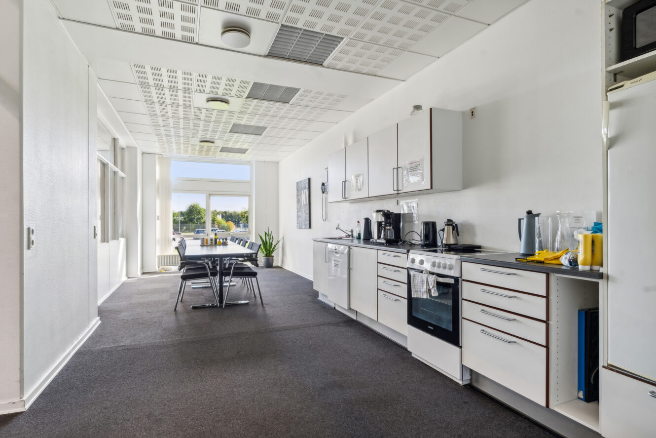 Et moderne kontorkøkken med hvide skabe, hvidevarer og et spisebord med stole nær store vinduer, der lukker naturligt lys ind.