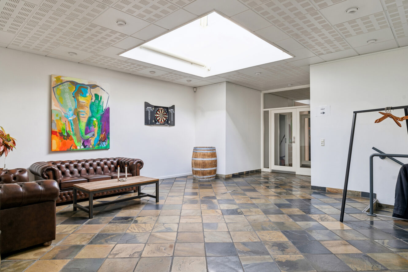 Et loungeområde med brune lædersofaer, et sofabord i træ, en dartskive på væggen, et abstrakt maleri, en tønde, en knagerække og et flisegulv under et ovenlysvindue.