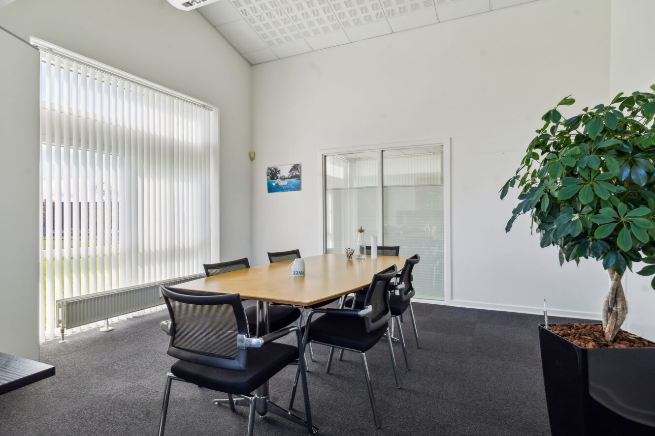 Et moderne konferencelokale med et træbord, seks sorte stole, store persienner, en potteplante og en skillevæg af glas.